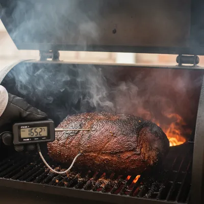 A person using a digital meat thermometer to check the internal temperature of food cooking on a pellet smoker.