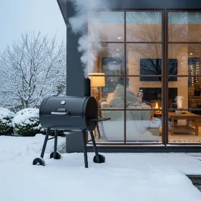 A heavy-duty pellet smoker operating in a snowy backyard with a user checking temperatures on a smartphone app indoors.
